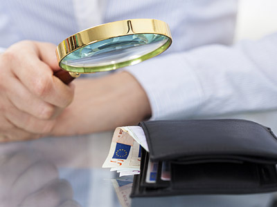 Eine Hand hält eine Lupe über einen Geldbeutel mit Euro-Scheinen.