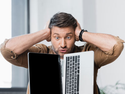 Ein Mann sitzt mit herausgestreckten Händen über dem Kopf vor einem Laptop und sieht überrascht oder gestresst aus.